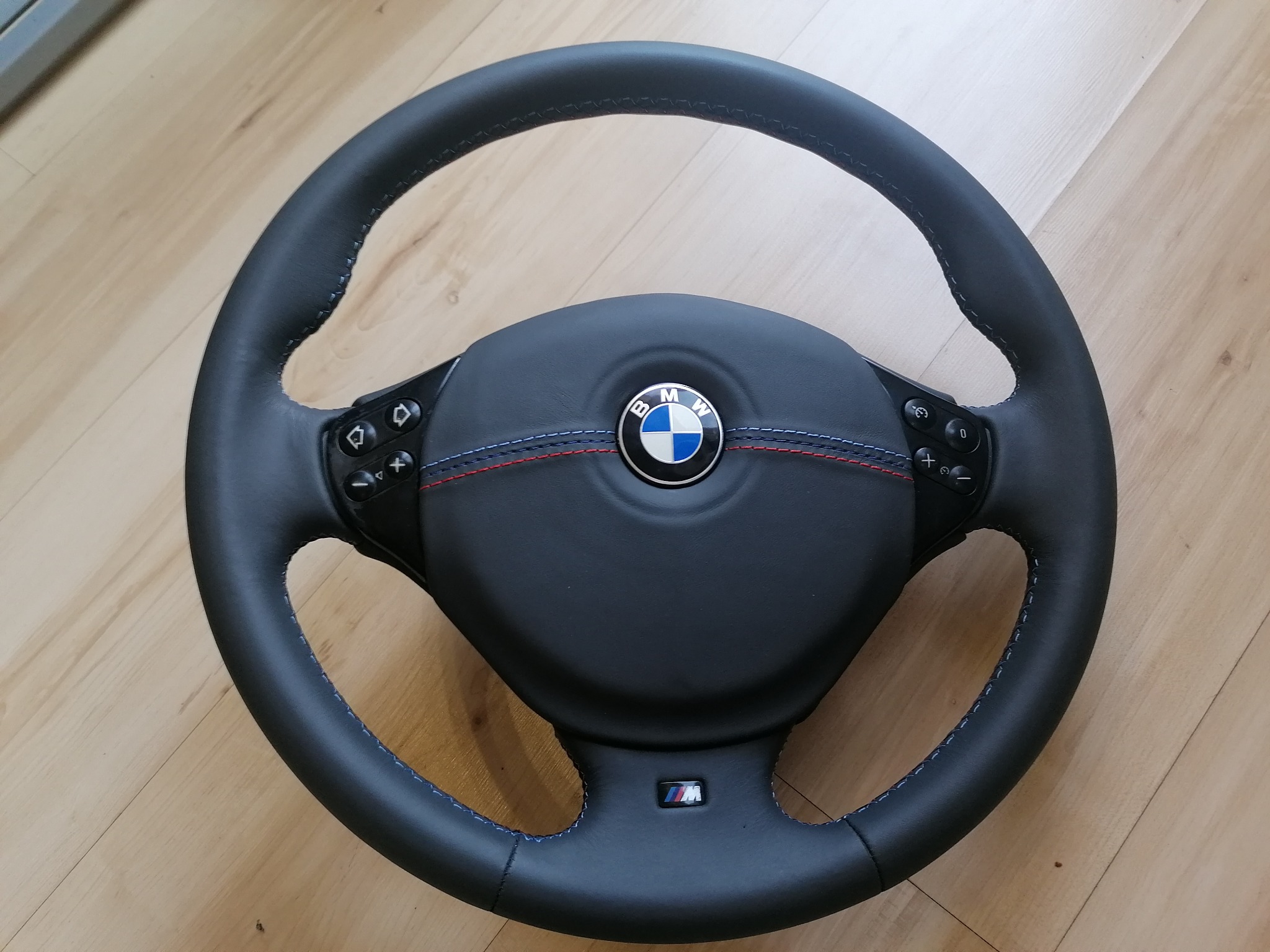 Odnowiona, czarna kierownica BMW M z precyzyjnym, niebieskim szwem, widoczna na drewnianym podłożu. Centralnie umieszczone logo BMW.