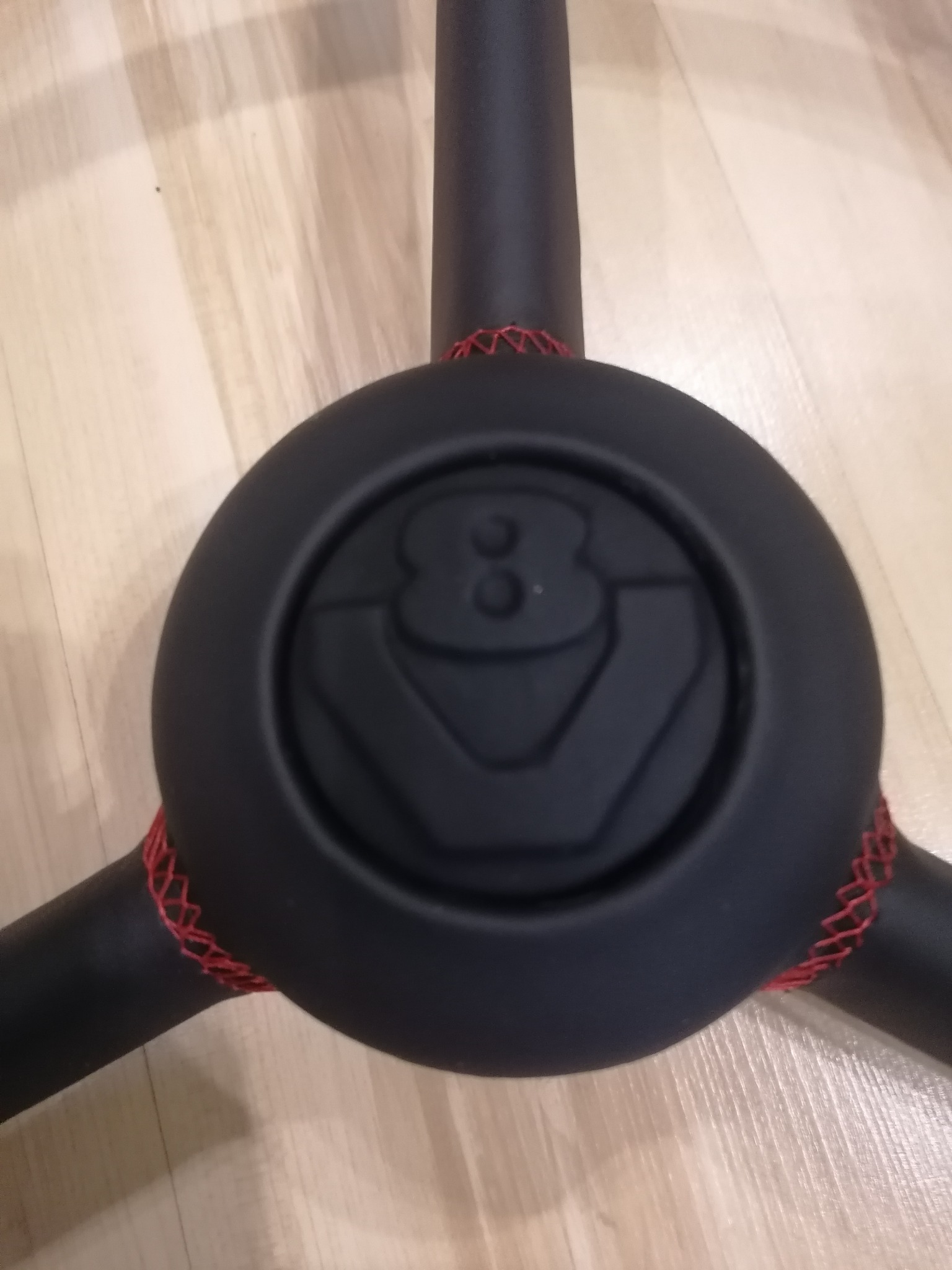 Czarny element fotela z logo i czerwonym szwem na drewnianej podłodze. Detal tapicerki samochodowej.