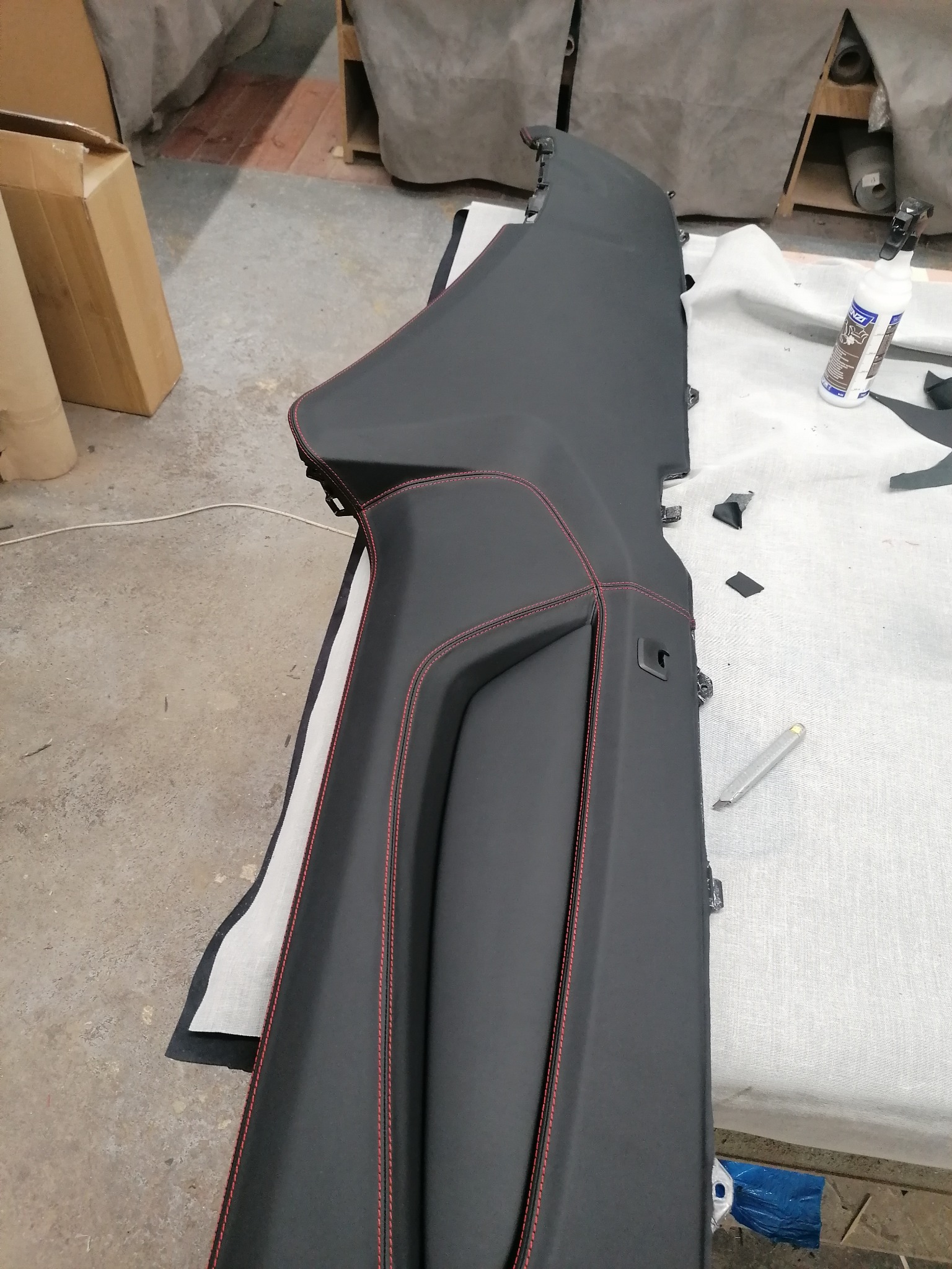 Czarny, tapicerowany element wnętrza auta z czerwonym szwem na stole warsztatowym. Widoczny nóż tapicerski i butelka kleju.