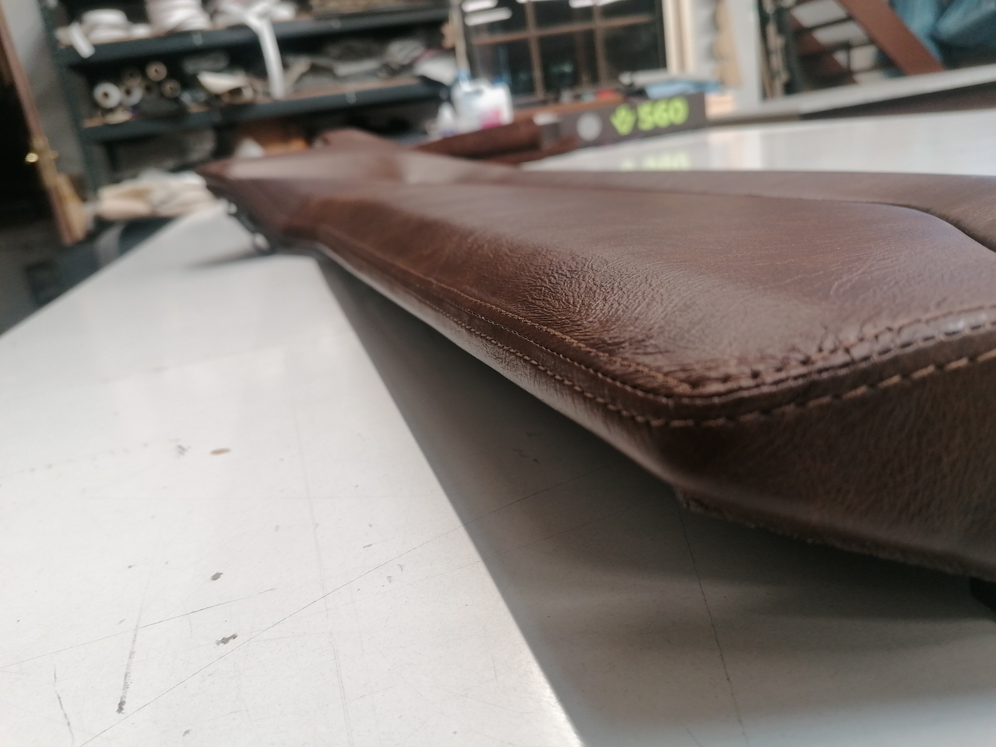 Brązowy, skórzany element tapicerowany z wyraźnym szwem na blacie roboczym, w tle warsztat tapicerski z półkami i narzędziami.