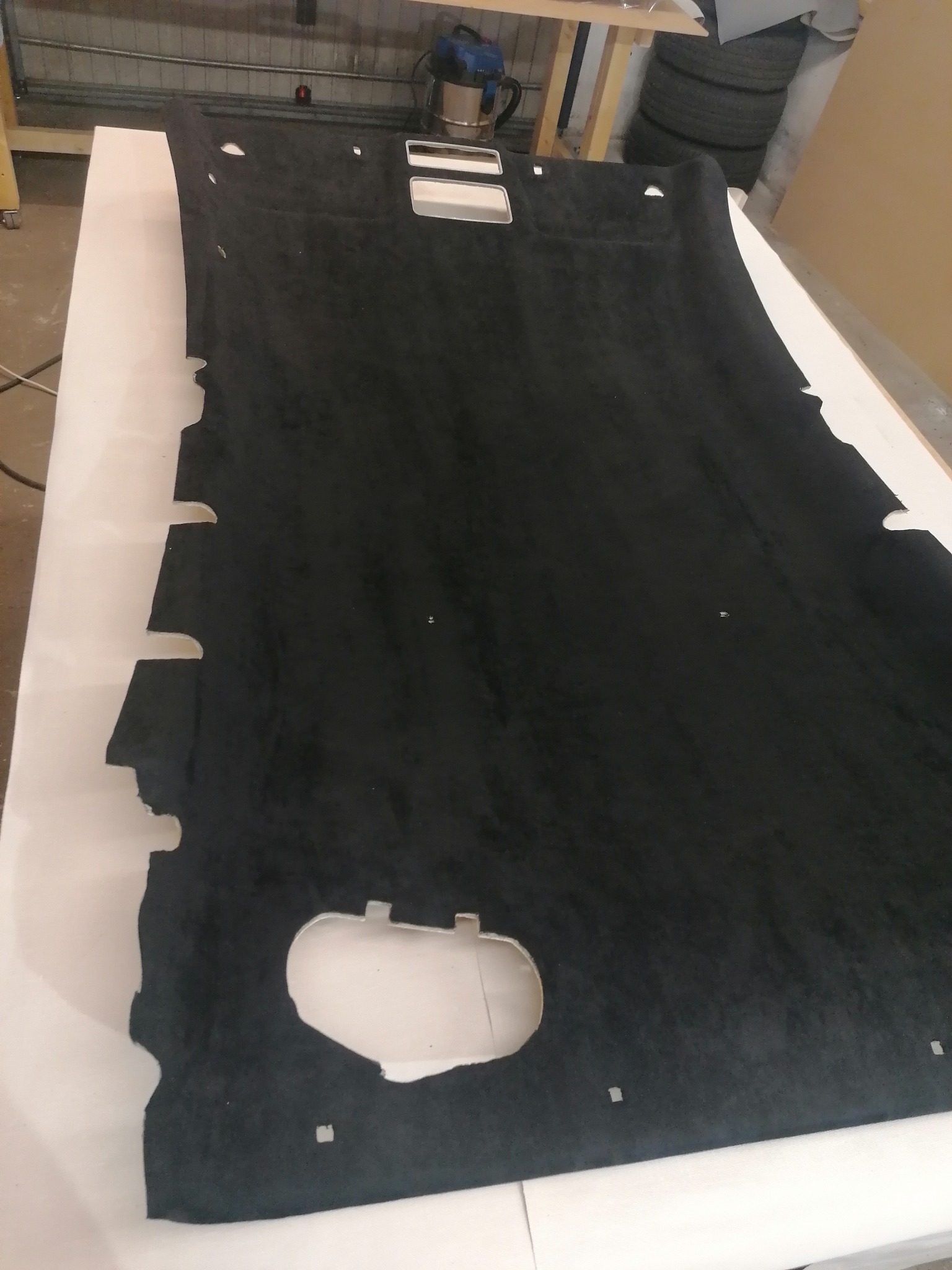 Czarna podsufitka samochodowa z wycięciami, leżąca na białym stole w warsztacie tapicerskim. Widoczne otwory na lampki i inne elementy wyposażenia.