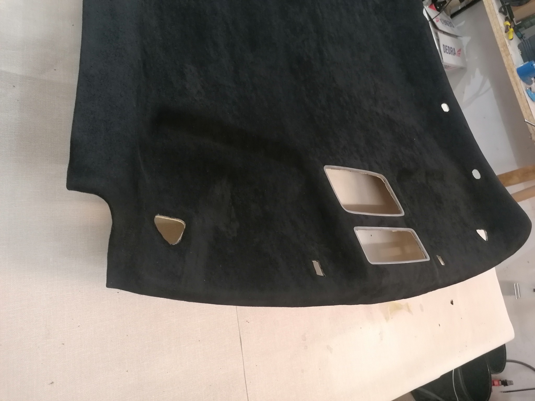 Czarna podsufitka samochodowa z wyciętymi otworami na oświetlenie i uchwyty, leżąca na stole w warsztacie. Widoczne narzędzia i akcesoria.