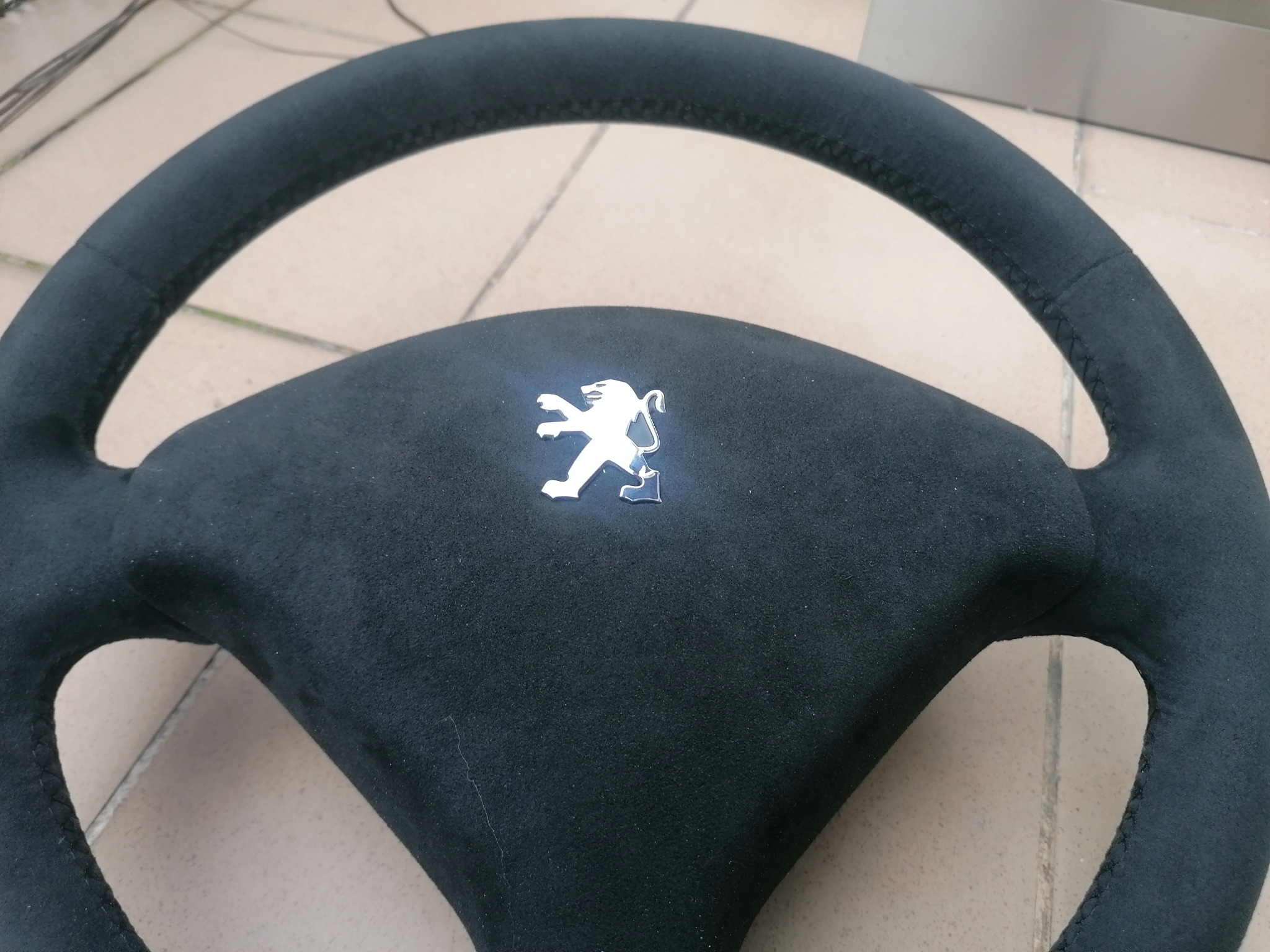 Obszyta czarną alcantarą kierownica Peugeot z logo lwa, widoczne szwy, leżąca na jasnej powierzchni. Detal tapicerki samochodowej po renowacji.