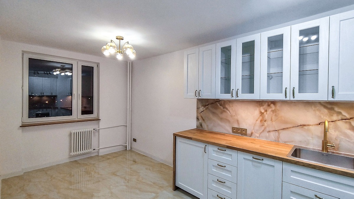 Jasna kuchnia z białymi szafkami, drewnianym blatem i marmurowym backsplashiem. Widoczne okno z odbiciem wnętrza. Złota bateria przy zlewie.