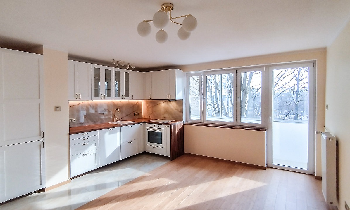 Jasna kuchnia z białymi szafkami, drewnianym blatem i marmurowym backsplashiem. Widok na okno z widokiem na drzewa. Nowoczesna lampa sufitowa.