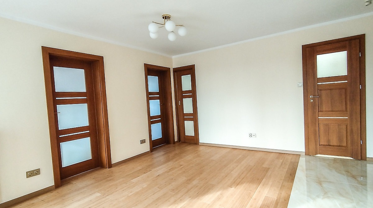 Jasne wnętrze z drewnianą podłogą i trzema drzwiami z przeszkleniami. Na suficie widoczna lampa z białymi kulami. Minimalistyczny design.