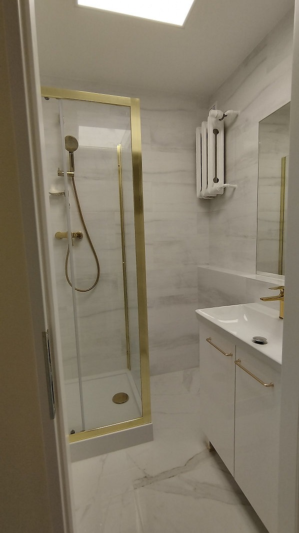 Elegancka łazienka z kabiną prysznicową w złotej ramie, białą umywalką z szafką i marmurowymi płytkami. Grzejnik drabinkowy na ścianie.
