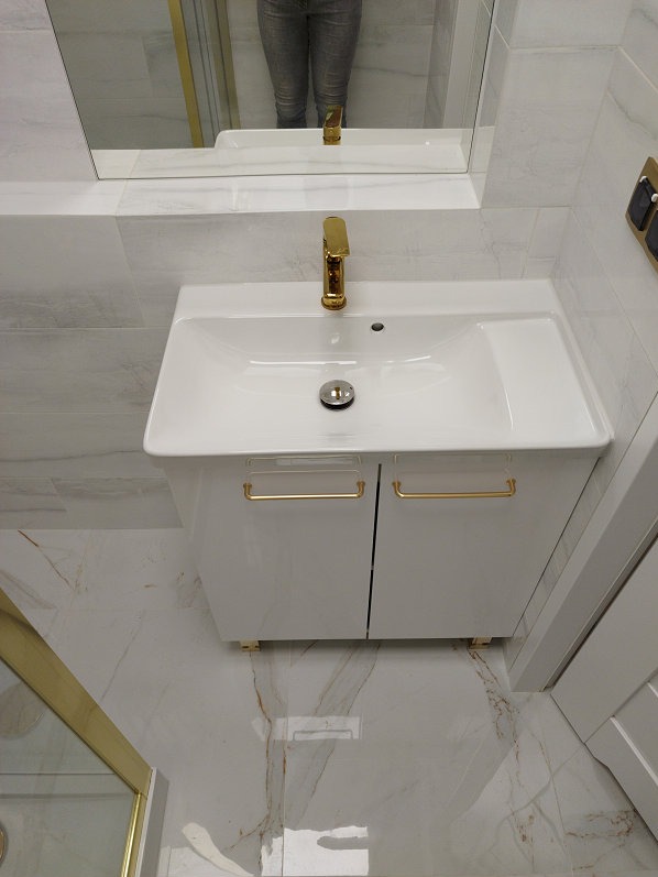 Biała umywalka z szafką i złotą armaturą w wykończonej łazience. W tle lustro z odbiciem osoby. Marmurowe płytki na podłodze i ścianach.