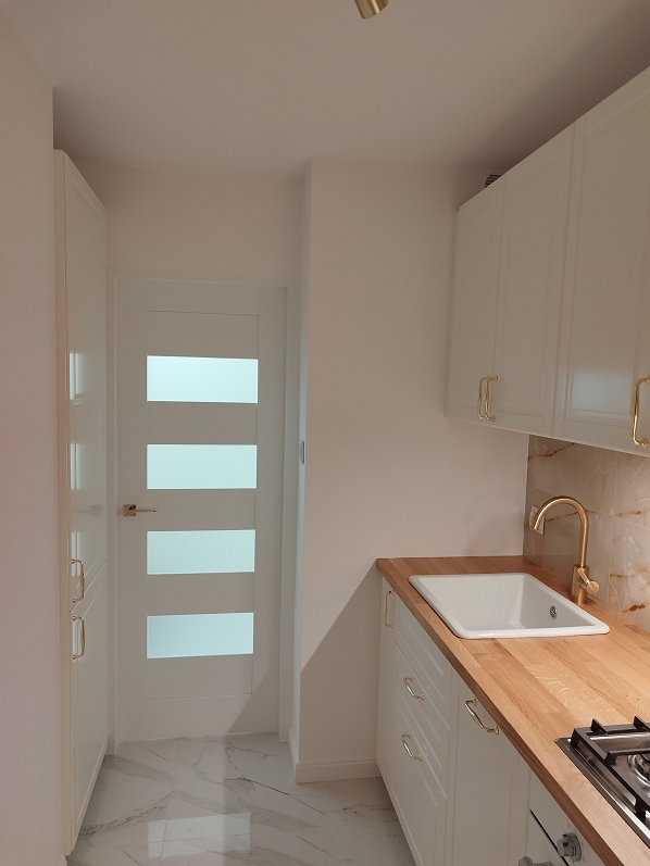 Jasna kuchnia z białymi szafkami, drewnianym blatem i złotą armaturą. Na podłodze marmurowe płytki, a obok drzwi z przeszkleniami.