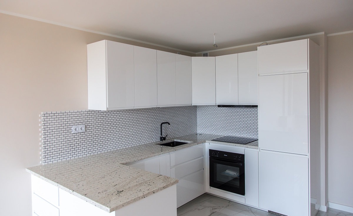 Biała, lśniąca kuchnia w nowoczesnym stylu z mozaiką na ścianie i blatem z jasnego granitu. Minimalistyczny design z czarną armaturą i piekarnikiem.