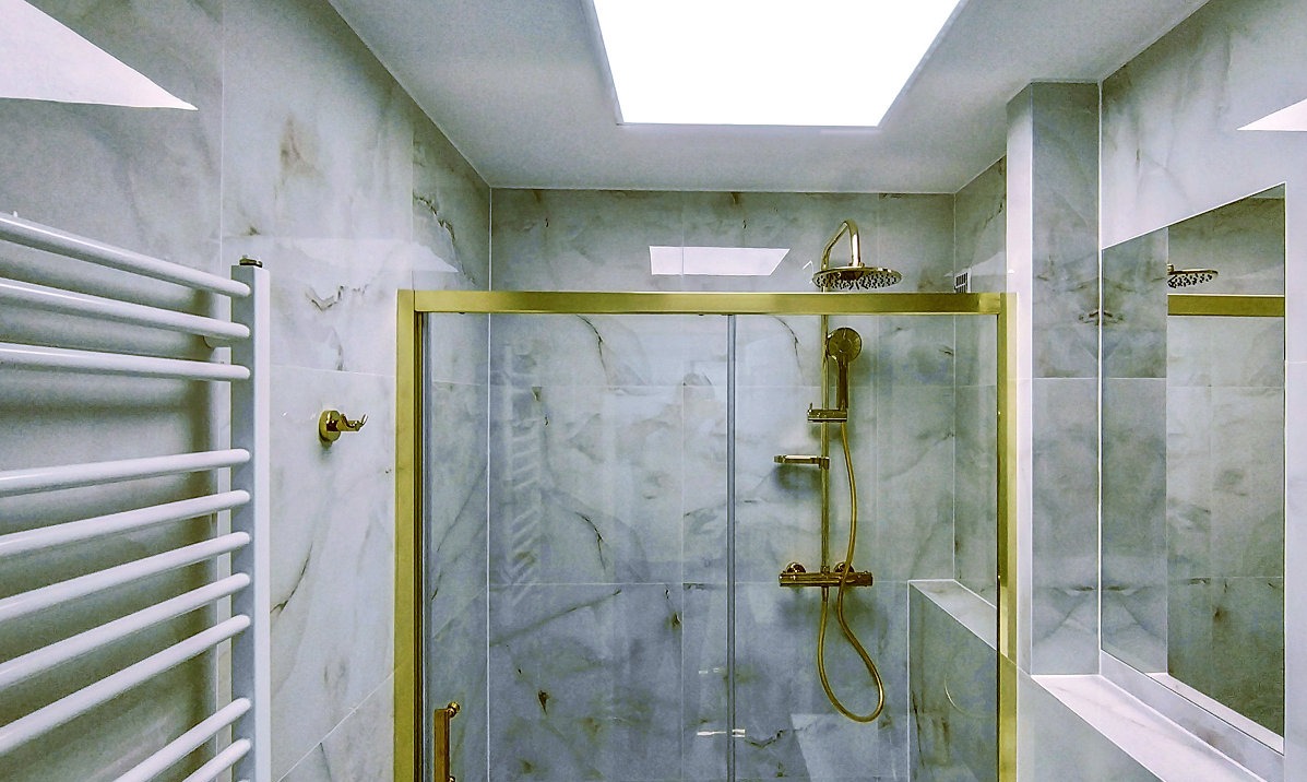 Elegancka łazienka z marmurowymi płytkami, złotą armaturą prysznicową i grzejnikiem drabinkowym. Sufit podświetlany. Lustro odbija fragment kabiny.