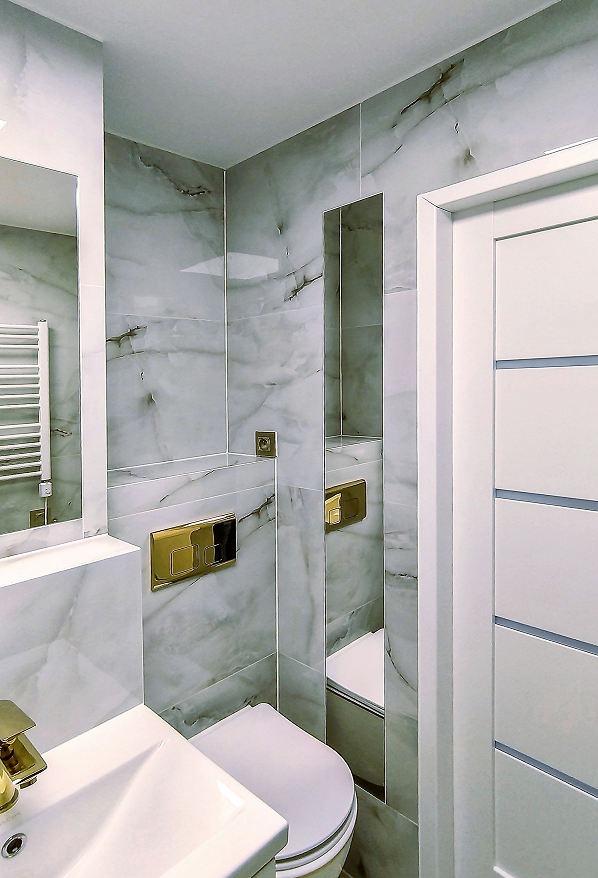 Jasna łazienka z marmurowymi płytkami, złotą armaturą i wnęką lustrzaną. Nowoczesne wykończenie, biała ceramika i drzwi z subtelnymi, szarymi detalami.