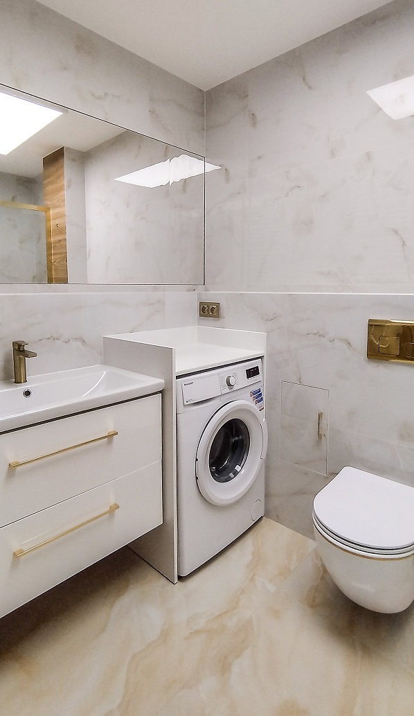 Jasna łazienka z marmurowymi płytkami, białą szafką z umywalką i złotymi uchwytami, pralką Sharp i podwieszaną toaletą.