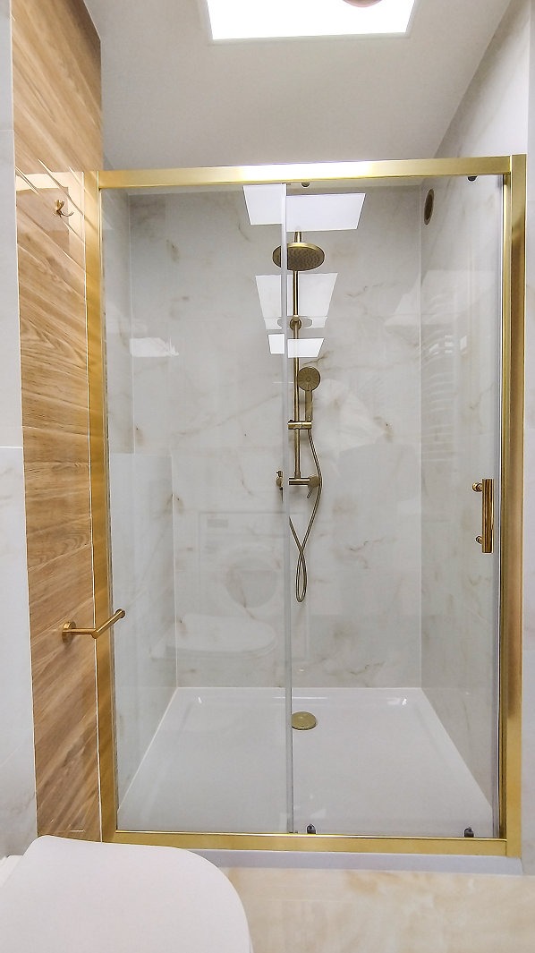 Kabina prysznicowa z przesuwnymi drzwiami w złotej ramie, marmurowe ściany, drewnopodobne płytki, widoczny fragment toalety i sufitowe okno.