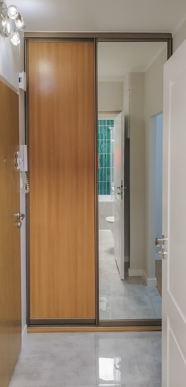 Szafa wnękowa z lustrem w przedpokoju, drewniane drzwi przesuwne, szara podłoga. Widok na łazienkę przez lustrzane drzwi szafy. Oświetlenie sufitowe.