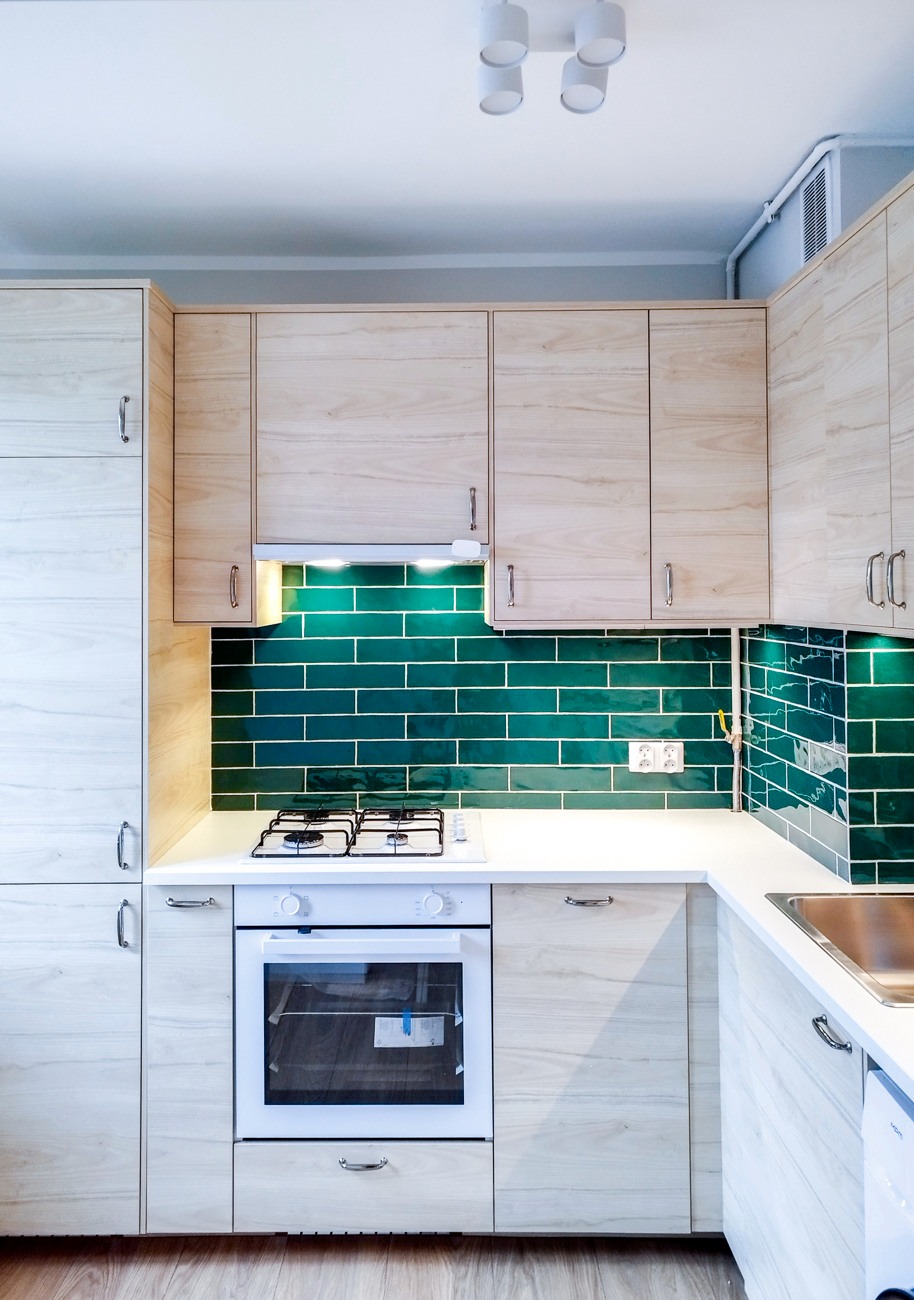 Jasna kuchnia z drewnianymi szafkami i zielonymi, błyszczącymi płytkami. Biały blat i sprzęt AGD. Nowoczesne oświetlenie sufitowe.