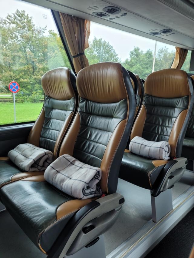 Wnętrze autobusu z brązowo-czarnymi skórzanymi fotelami i kocami w kratę. Widok przez okno na zieleń i znak drogowy.