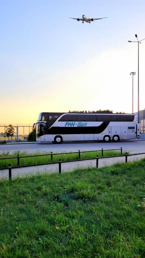 Czarno-biały, dwupiętrowy autokar PAN-BUS na tle lotniska i startującego samolotu. Ujęcie z dołu, w tle zachodzące słońce.