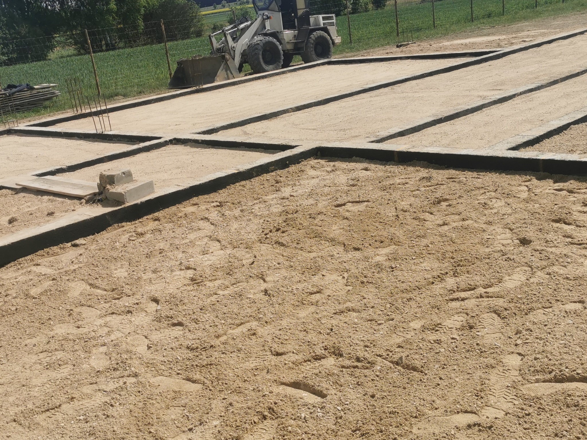 Zalany piaskiem fundament z betonowymi belkami, widoczny z bliska, z ładowarką w tle. Ziemia ubita, ślady opon. Widok na zielone pole i drzewa w oddali.