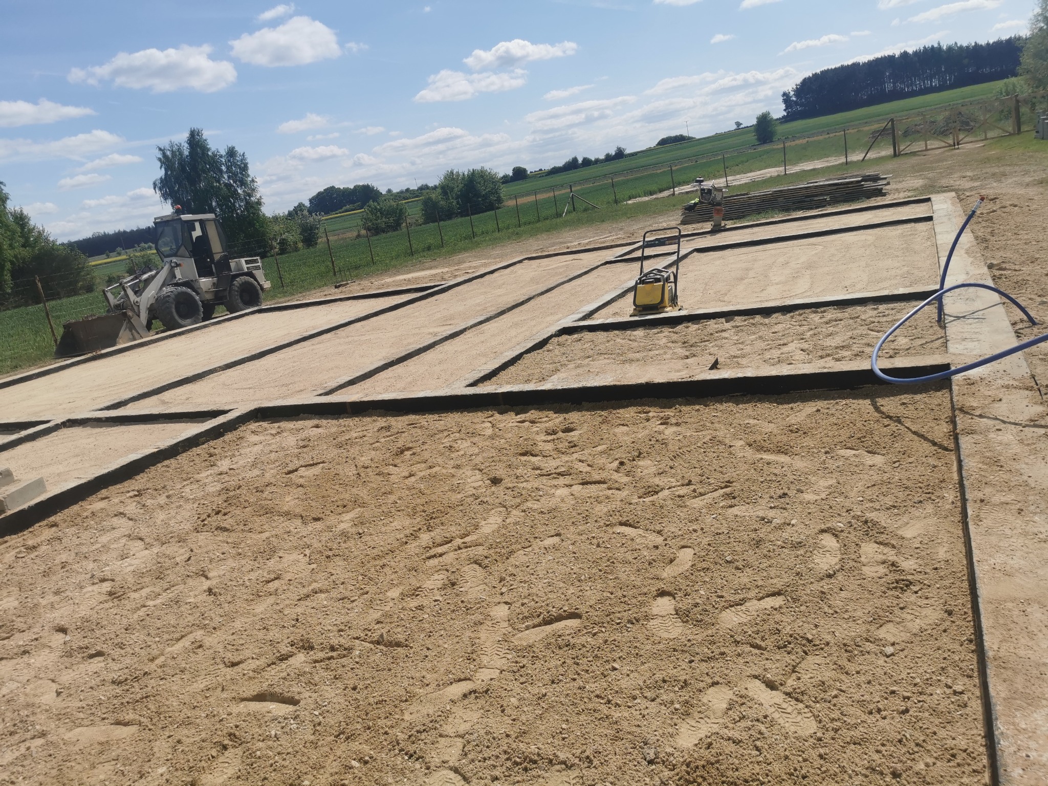 Przygotowanie terenu pod fundamenty: wytyczone obrysy, ubijarka i miniładowarka na placu budowy. Widoczne ślady stóp na piasku i częściowo ułożone deski.