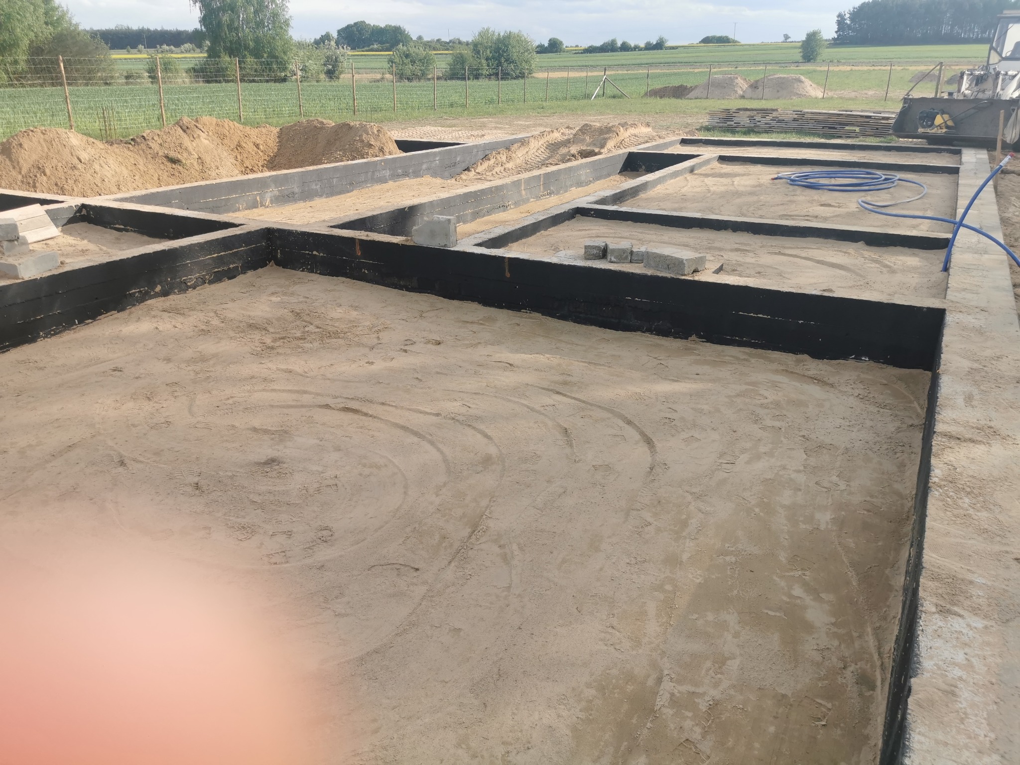 Zabezpieczone fundamenty pod budowę domu jednorodzinnego, widoczne szalunki i przygotowany teren. W tle krajobraz polny i sprzęt budowlany.