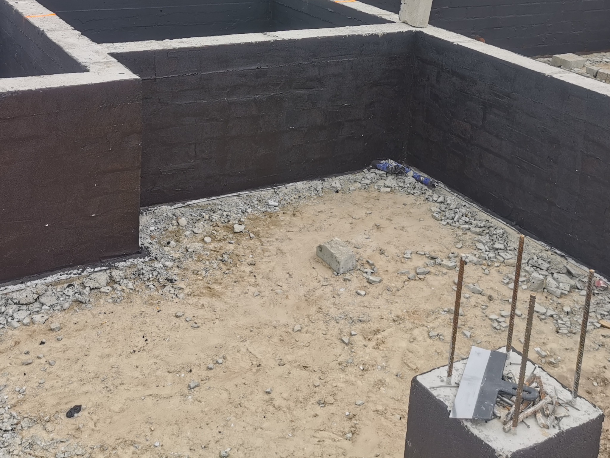 Zabezpieczone fundamenty domu, izolacja przeciwwilgociowa ścian fundamentowych, teren przygotowany pod dalsze prace. Widoczne pręty zbrojeniowe i narzędzia.