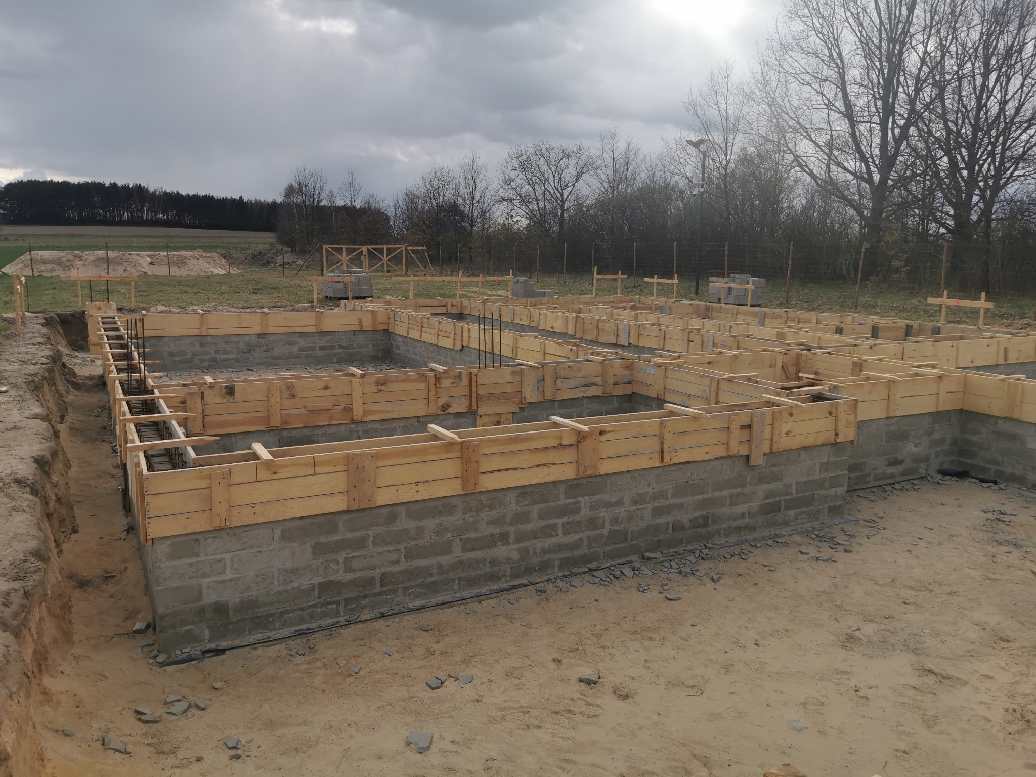 Fundamenty domu jednorodzinnego w trakcie budowy: wylane ławy fundamentowe z szalunkiem drewnianym, zbrojenie pionowe. Na drugim planie drzewa i pochmurne niebo.