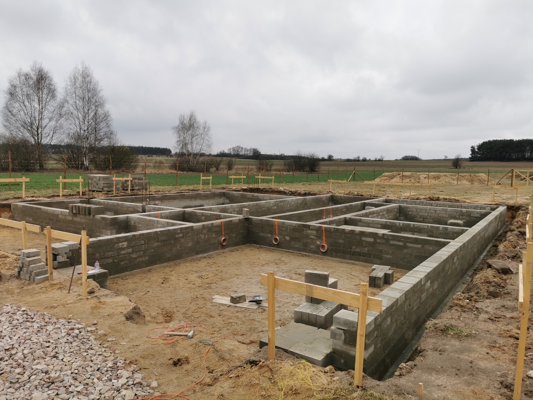 Wykopane i wymurowane fundamenty pod budowę domu jednorodzinnego. Widoczne bloczki betonowe, drewniane szalunki i teren budowy z polami uprawnymi w tle.