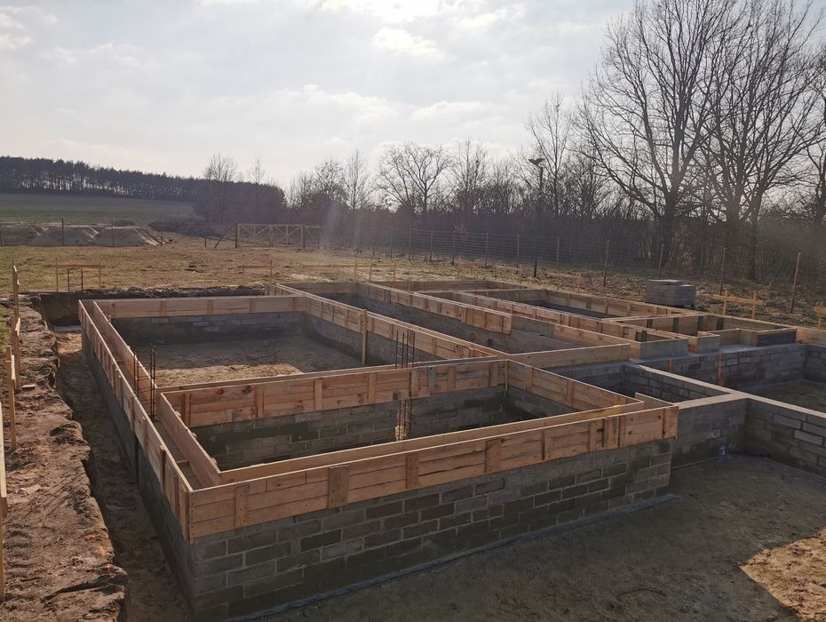 Zalane fundamenty pod budowę domu, widoczne szalunki i wystające pręty zbrojeniowe. W tle zalesiony teren i błękitne niebo.