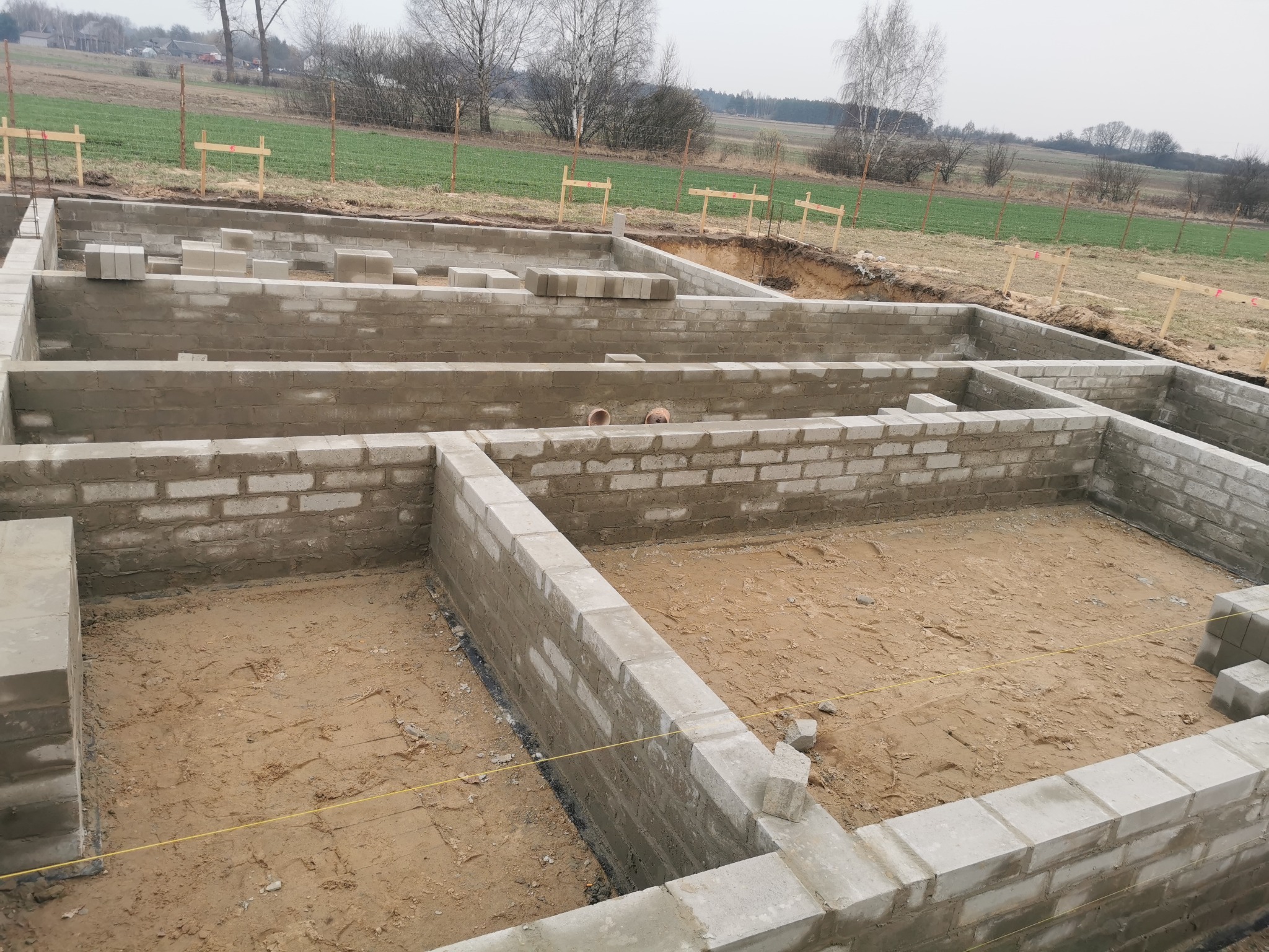 Fundamenty z bloczków betonowych, częściowo wymurowane, widok z góry na plac budowy z zarysem przyszłego budynku na tle drzew i pola.