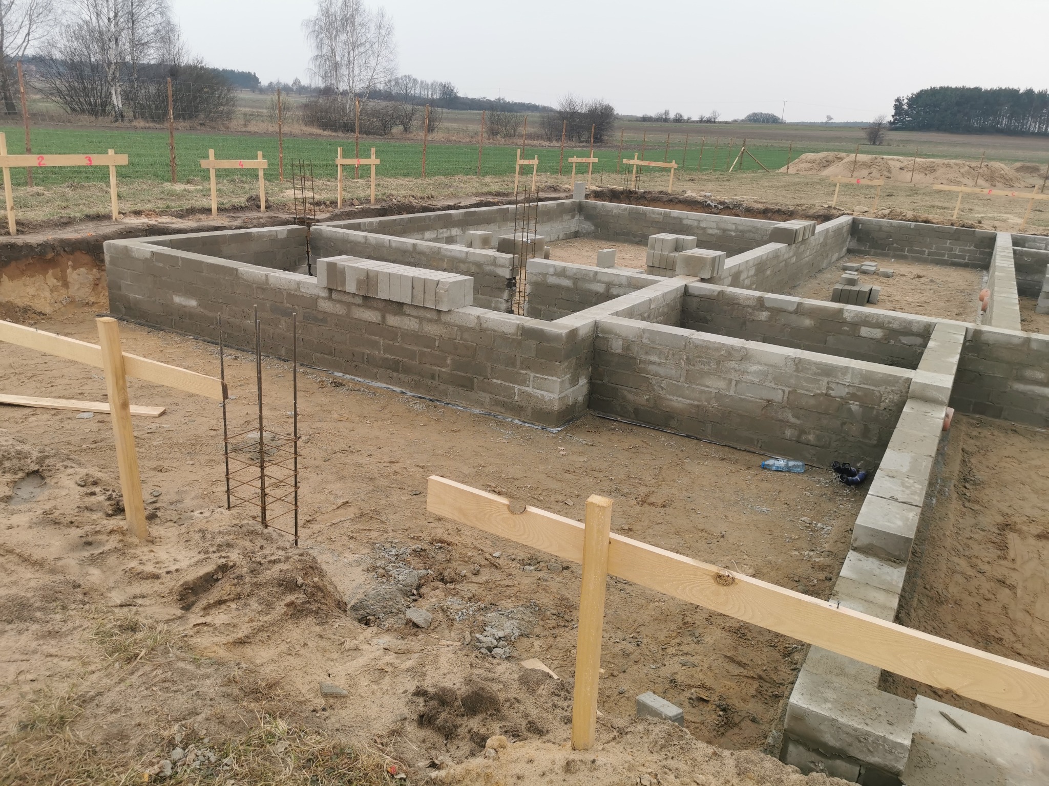 Fundamenty domu z betonu komórkowego w trakcie budowy, widoczne zbrojenia i drewniane oznaczenia. Tło stanowią pola i las.
