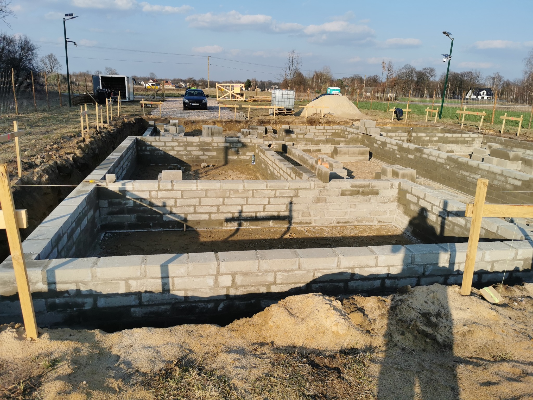 Fundamenty z bloczków betonowych na działce budowlanej, widoczne szalunki i wykop. W tle samochód i drzewa.