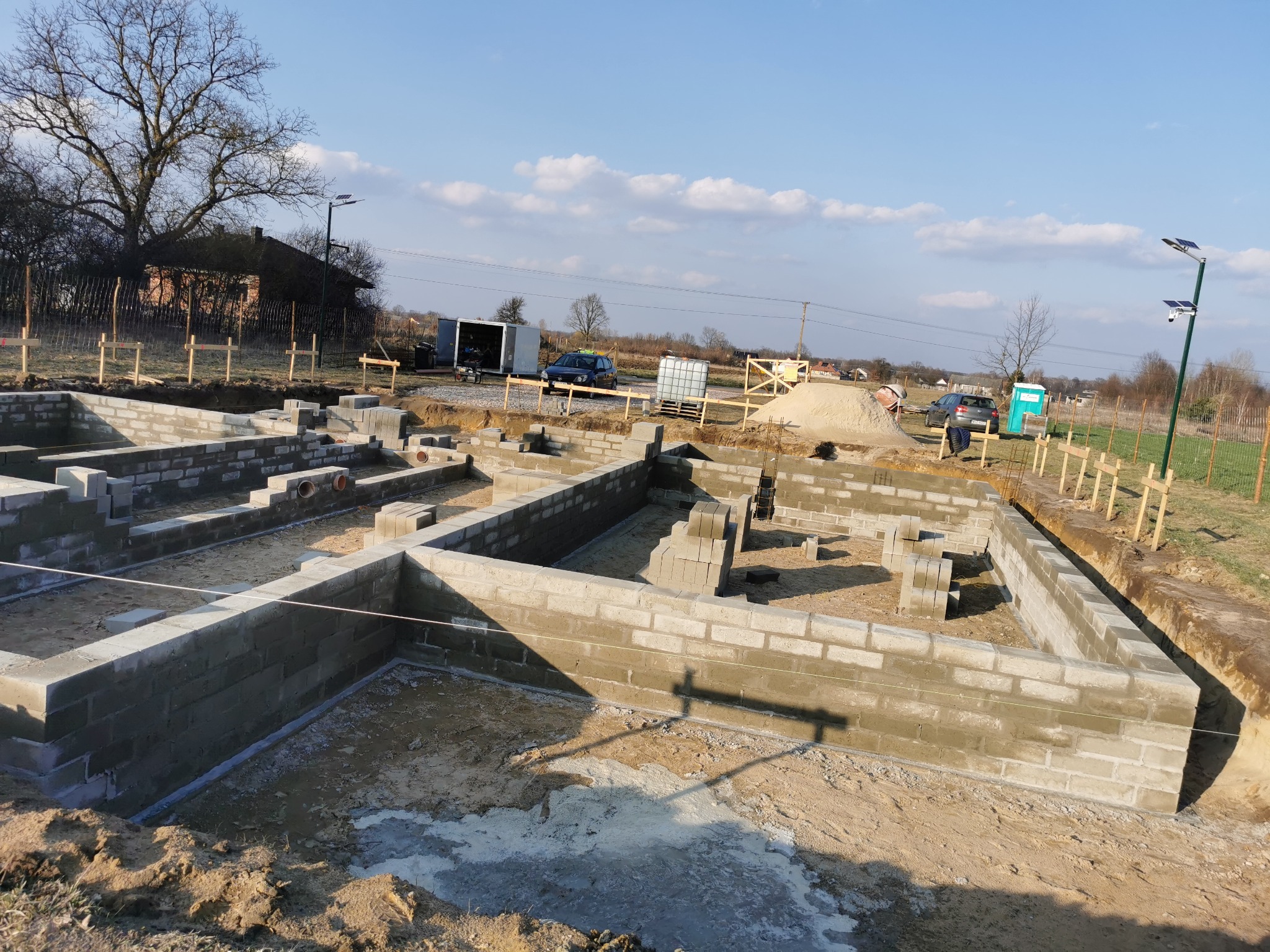 Wymurowane ściany fundamentowe z bloczków betonowych na działce budowlanej, widok z góry. Część bloczków jeszcze nieużyta. W tle drzewa, dom i elementy infrastruktury.