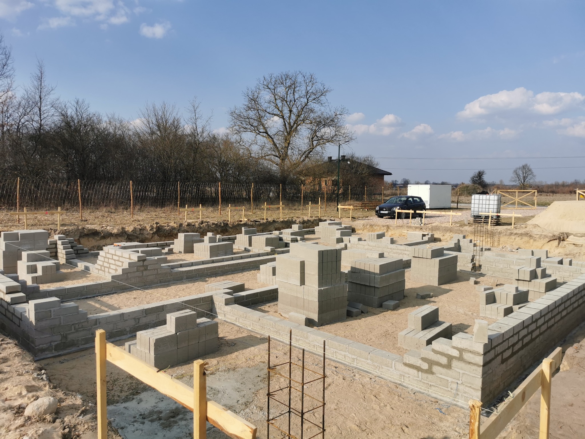 Fundamenty z bloczków betonowych na piaszczystym gruncie, widoczne zbrojenia, w tle drzewa i budynek w słoneczny dzień.