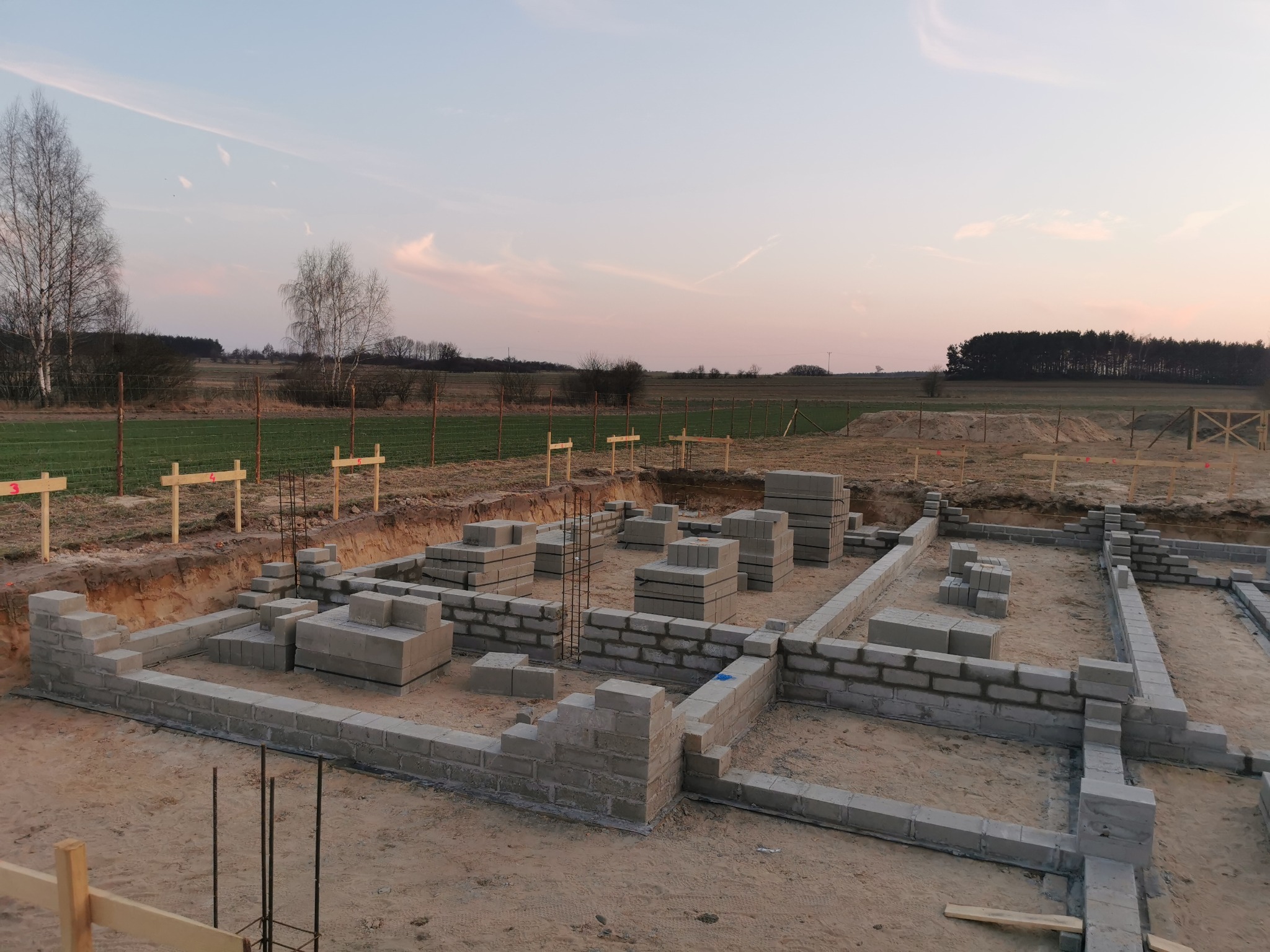 Fundamenty domu z bloczków betonowych na piaszczystym gruncie, zbrojenie widoczne w narożnikach. W tle pole i drzewa pod wieczornym niebem.