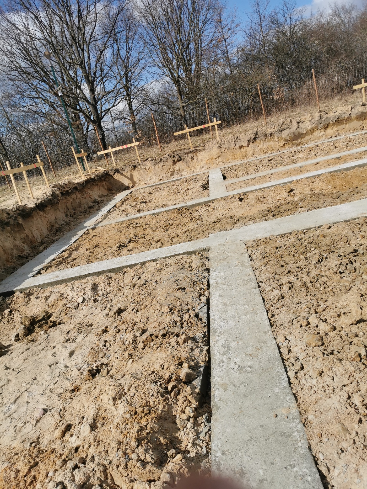 Zalane betonem ławy fundamentowe w wykopie, widoczne drewniane znaczniki. Prace ziemne i betonowe w słoneczny dzień. Elementy zbrojenia wystają z betonu.
