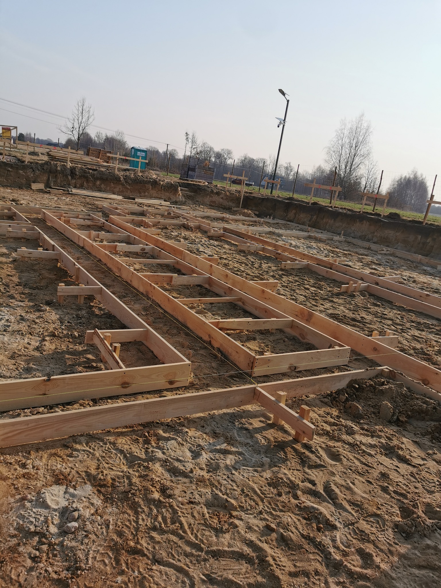 Drewniane szalunki ułożone w wykopie, przygotowane do zalania fundamentów. Widoczny grunt i elementy konstrukcyjne z drewna.