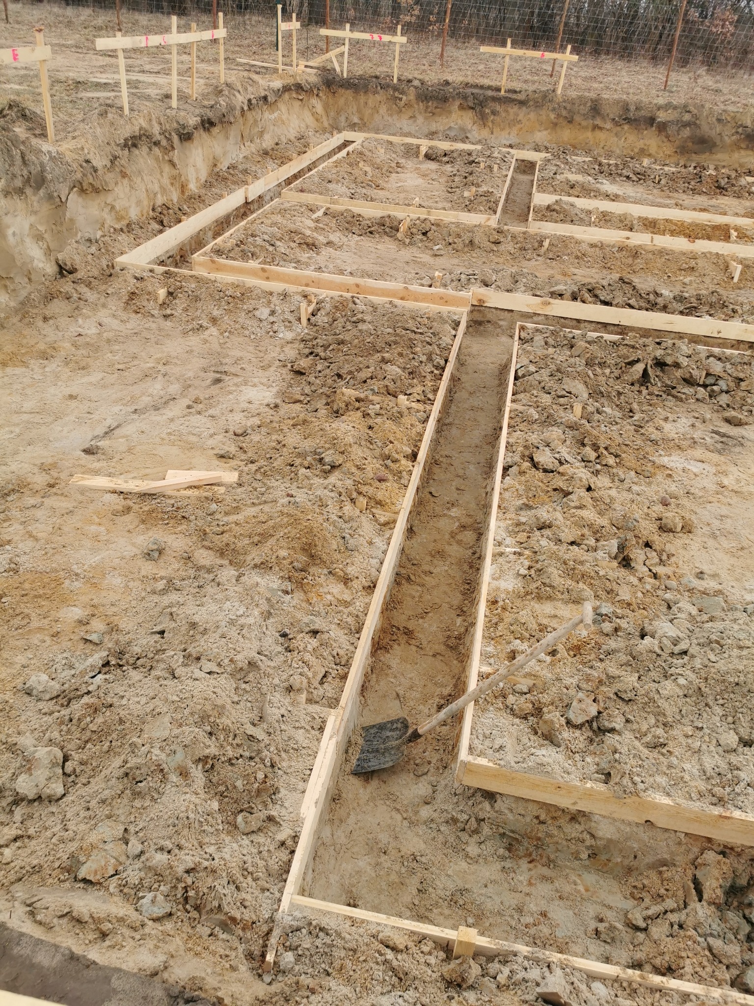 Szeroki kadr na wykop pod fundamenty z drewnianym szalunkiem. W wykopie leży łopata. W tle widoczne drewniane znaczniki i siatka ogrodzeniowa.