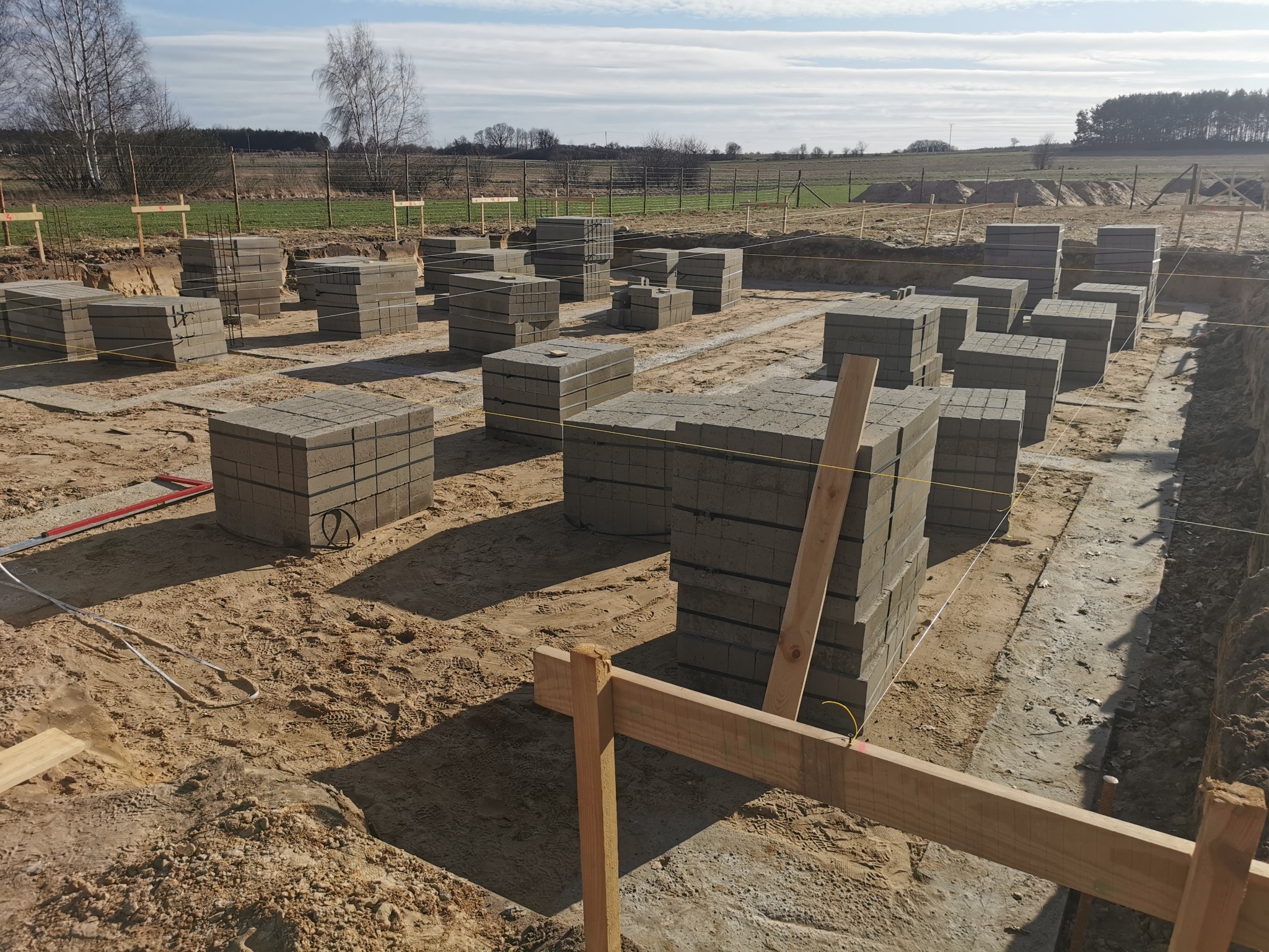 Fundamenty z bloczków betonowych na budowie domu, widok z góry. Ustawione w rzędach, teren przygotowany, drewniane szalunki w narożnikach.