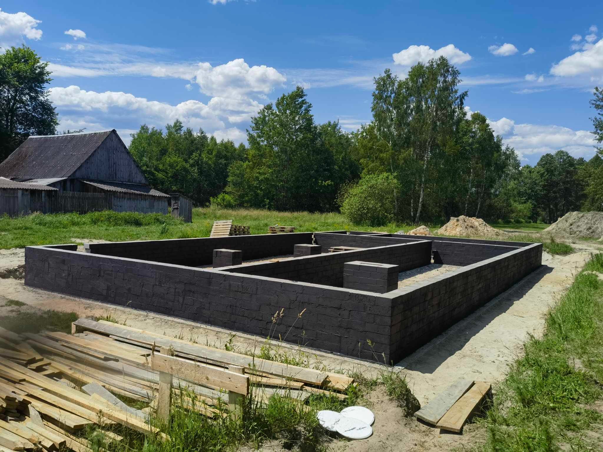 Czarny fundament z cegły na tle wiejskiego krajobrazu pod błękitnym niebem. Widoczne deski i roślinność w otoczeniu.