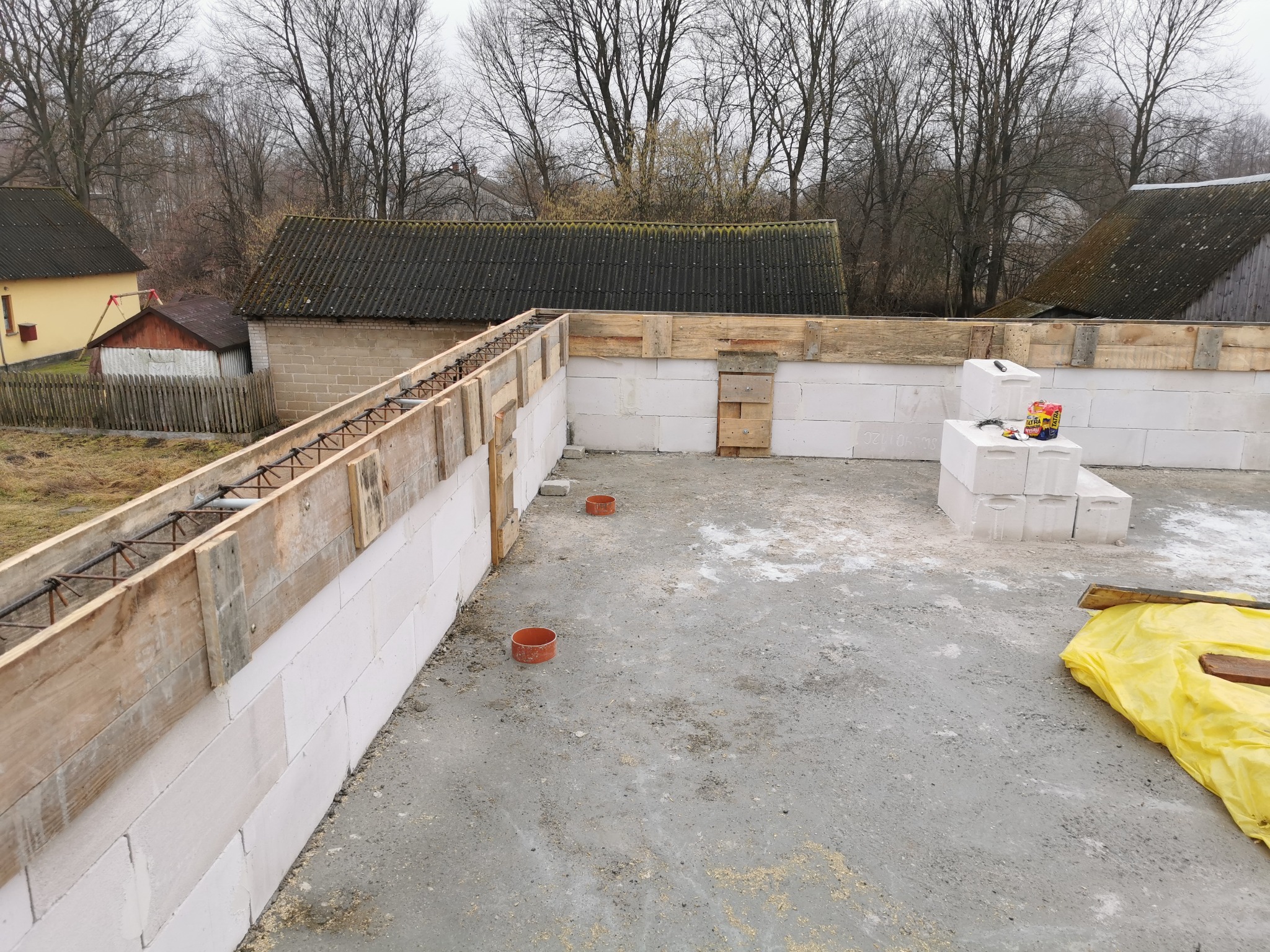 Wznoszenie ścian z bloczków betonu komórkowego z szalunkiem zbrojeniowym. Widoczne bloczki, szalunek, zbrojenie oraz fragment fundamentu. Prace budowlane w toku.