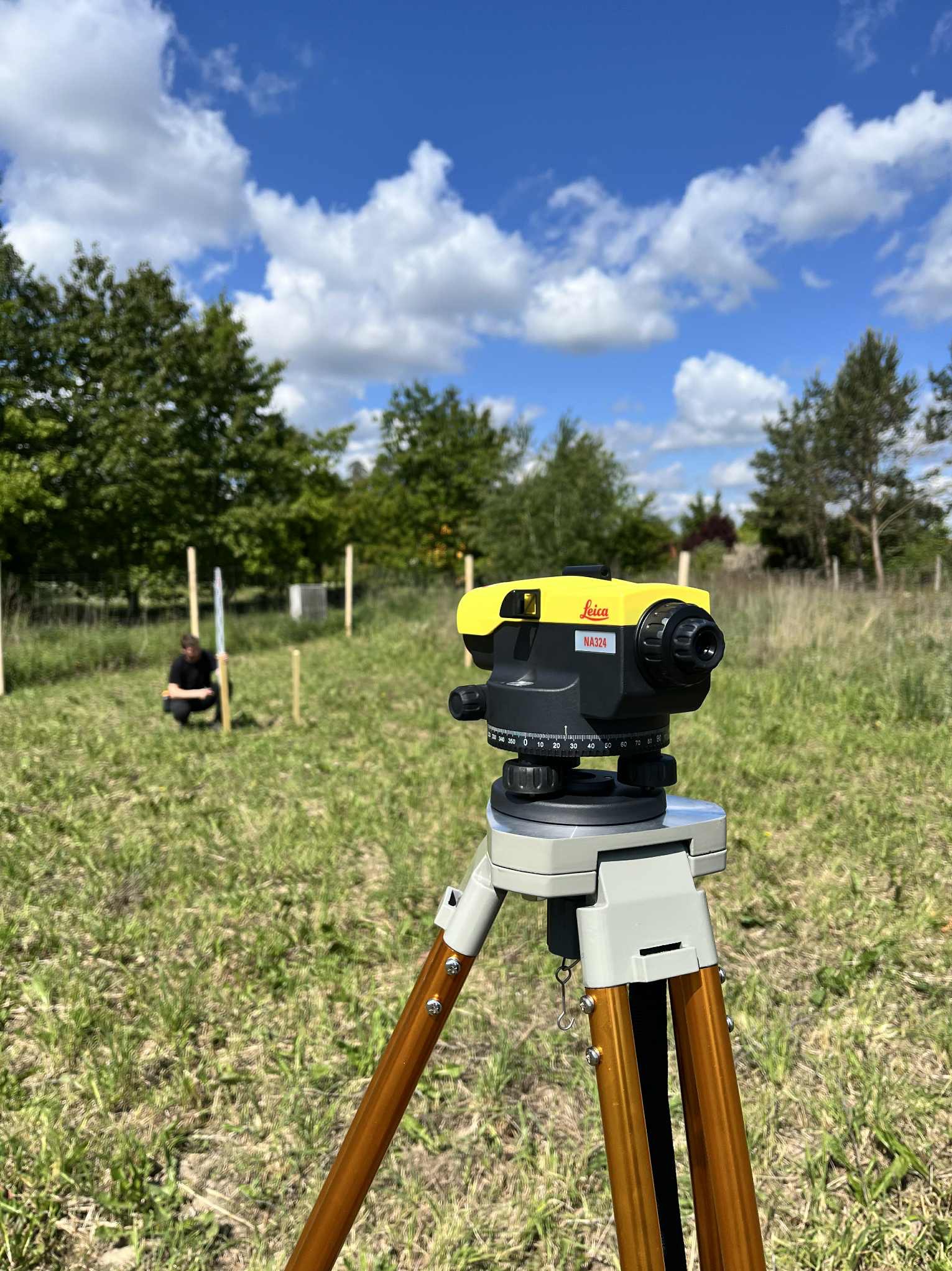 Żółty niwelator Leica NA324 na statywie, w tle osoba przy palikach na łące pod błękitnym niebem z chmurami. Prace pomiarowe w terenie.
