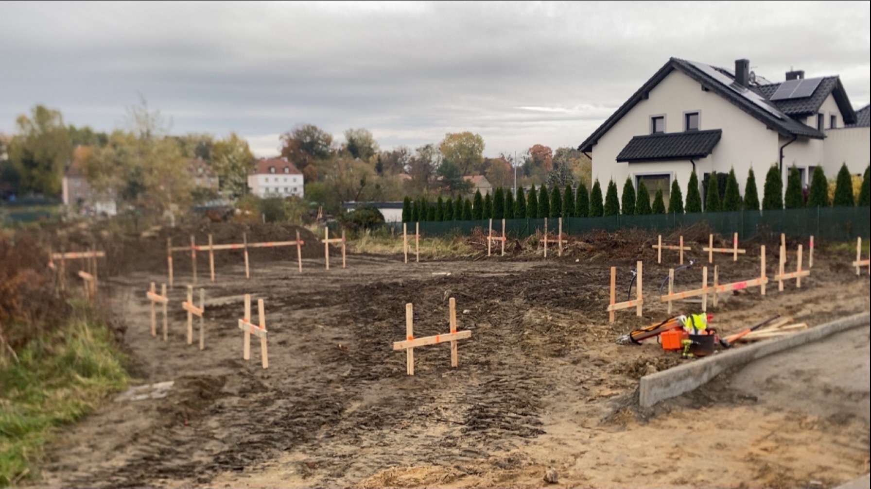 Wytyczenie fundamentów pod budowę domu jednorodzinnego. Widoczne drewniane znaczniki geodezyjne na działce, w tle zabudowa mieszkalna i pochmurne niebo.