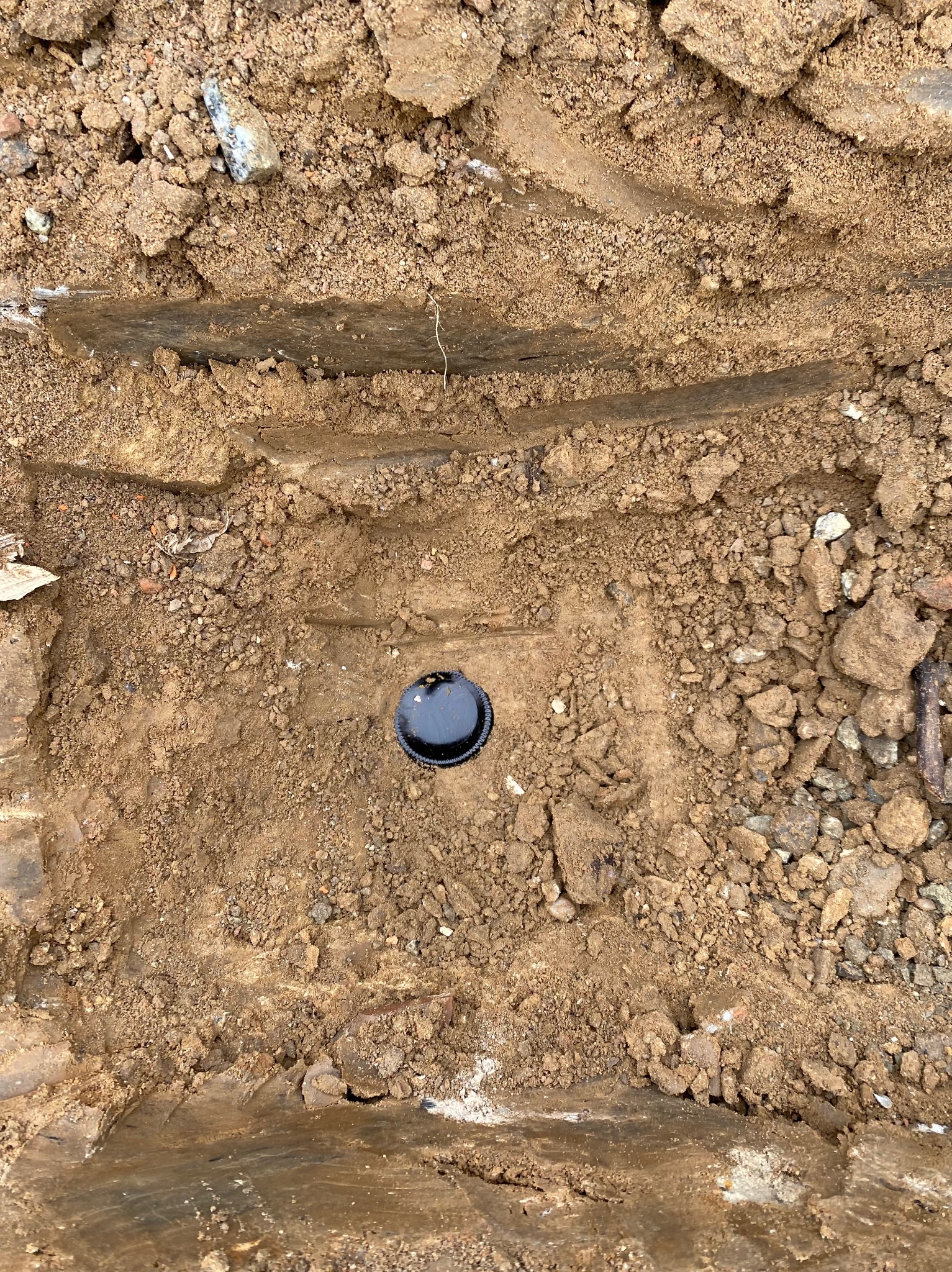 Odkrywka ziemna z widocznym czarnym włazem kanalizacyjnym. Ślady opon na ziemi. Prace ziemne w trakcie realizacji.