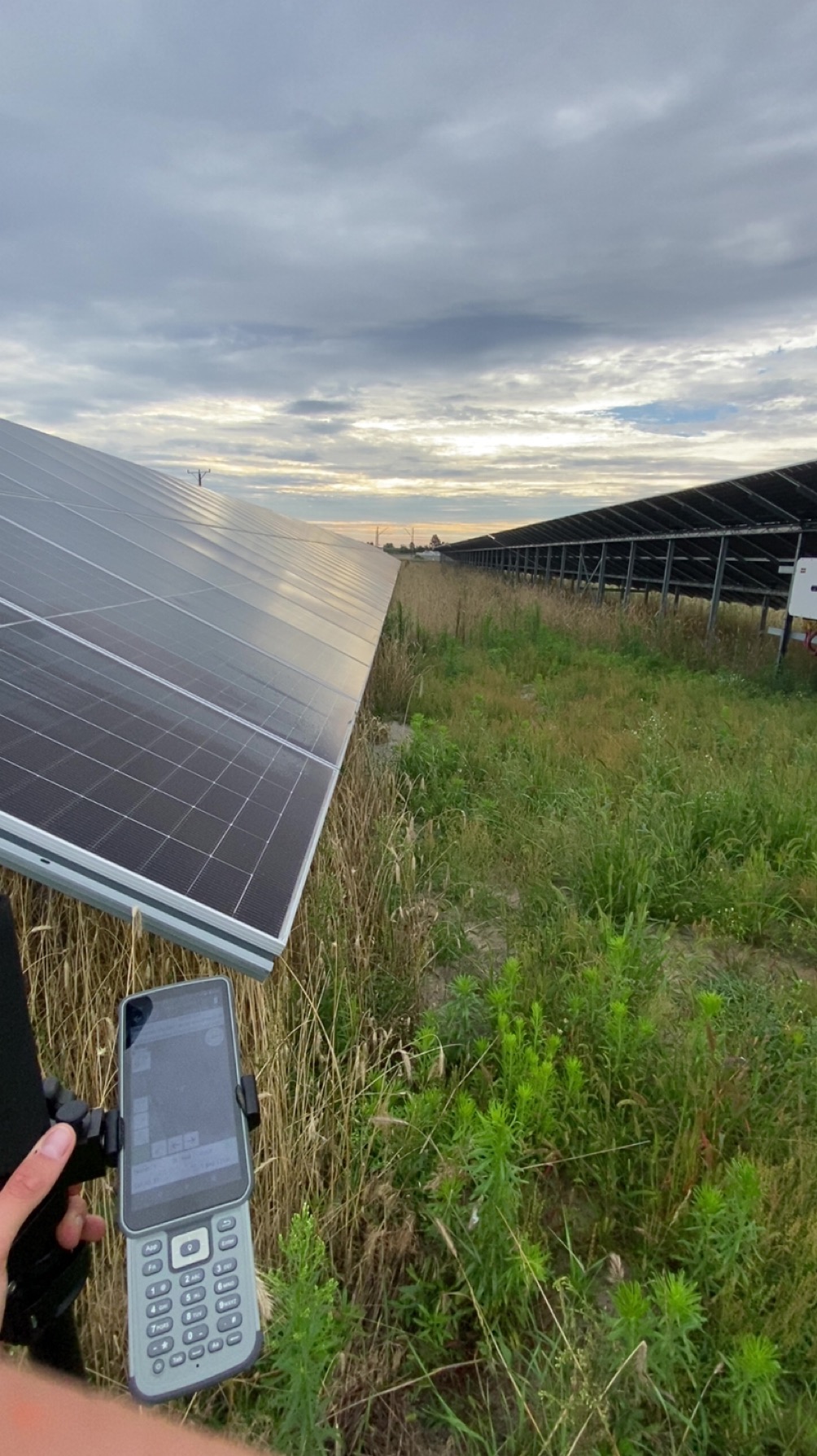 Geodezyjny pomiar paneli słonecznych we Wrześni. Urządzenie pomiarowe w dłoni na tle rzędów paneli i zachmurzonego nieba. Prace geodezyjne przy farmie fotowoltaicznej.