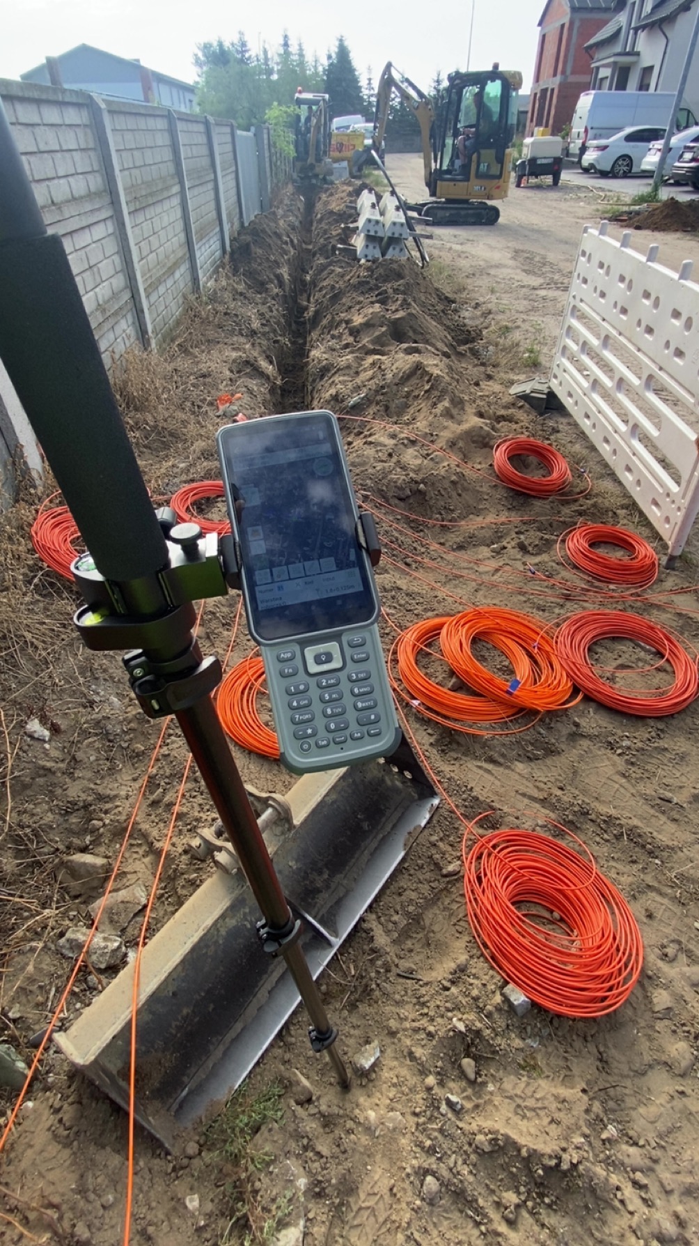 Prace ziemne z użyciem specjalistycznego sprzętu pomiarowego i pomarańczowych kabli. W tle wykop, maszyny budowlane i ogrodzenie.