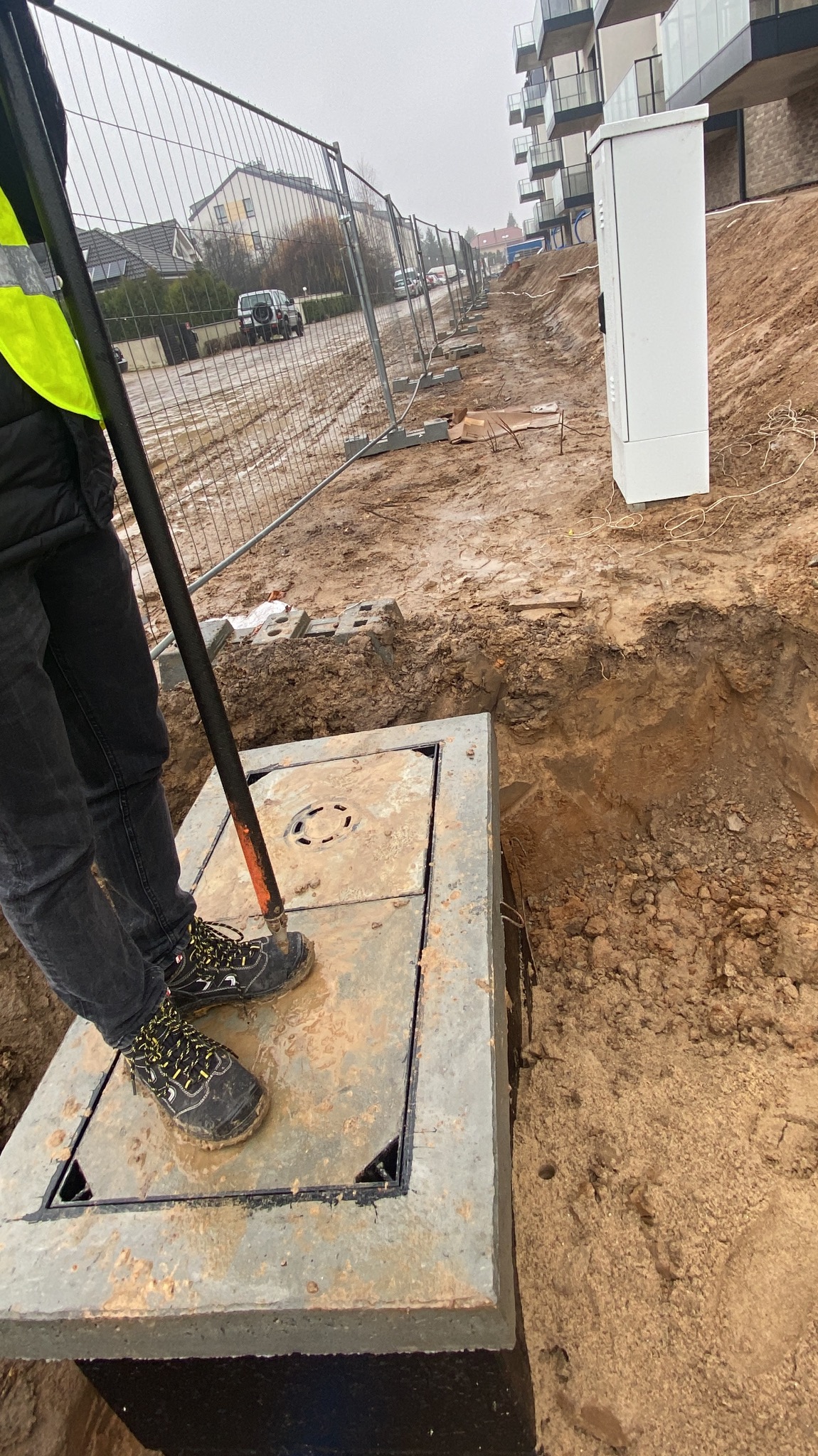 Pracownik w odblaskowej kamizelce stoi na betonowej studzience kanalizacyjnej na placu budowy, w tle ogrodzenie i budynki mieszkalne.