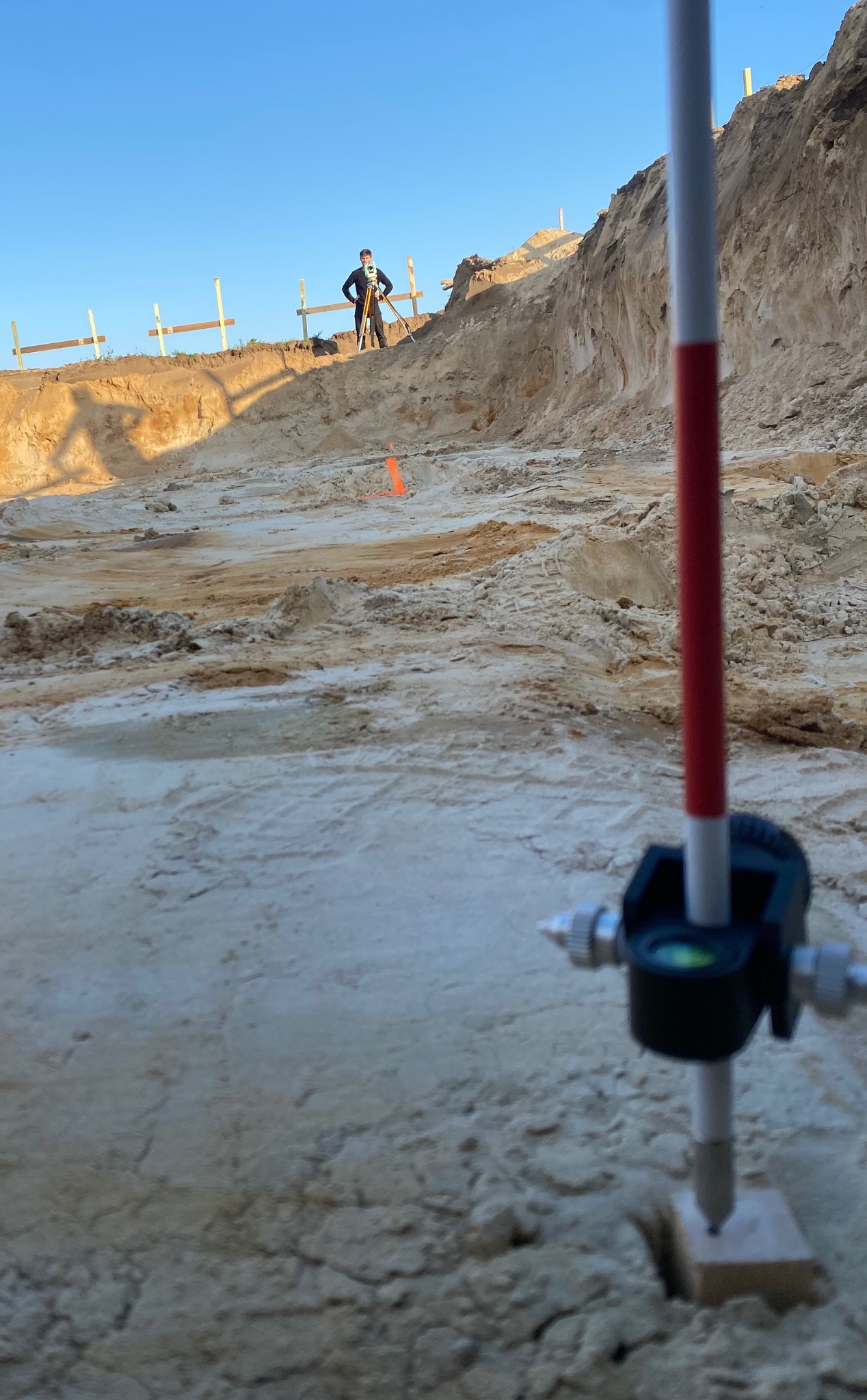 Prace pomiarowe na wykopie budowlanym. W tle widoczny pracownik z niwelatorem. W pierwszym planie tyczka geodezyjna z libellą.