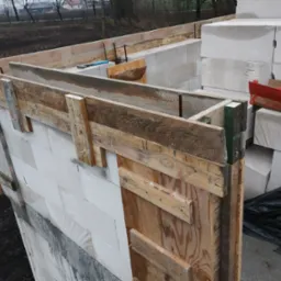 Budowa ściany z bloczków betonu komórkowego Ytong, z drewnianym szalunkiem i widocznymi narzędziami. W tle ziemia i zabudowania.