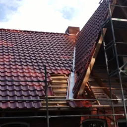 Częściowo zdjęte czerwone dach&oacute;wki na budynku z cegły, widoczne rusztowanie i elementy więźby. Etap wymiany pokrycia dachowego w słoneczny dzień.